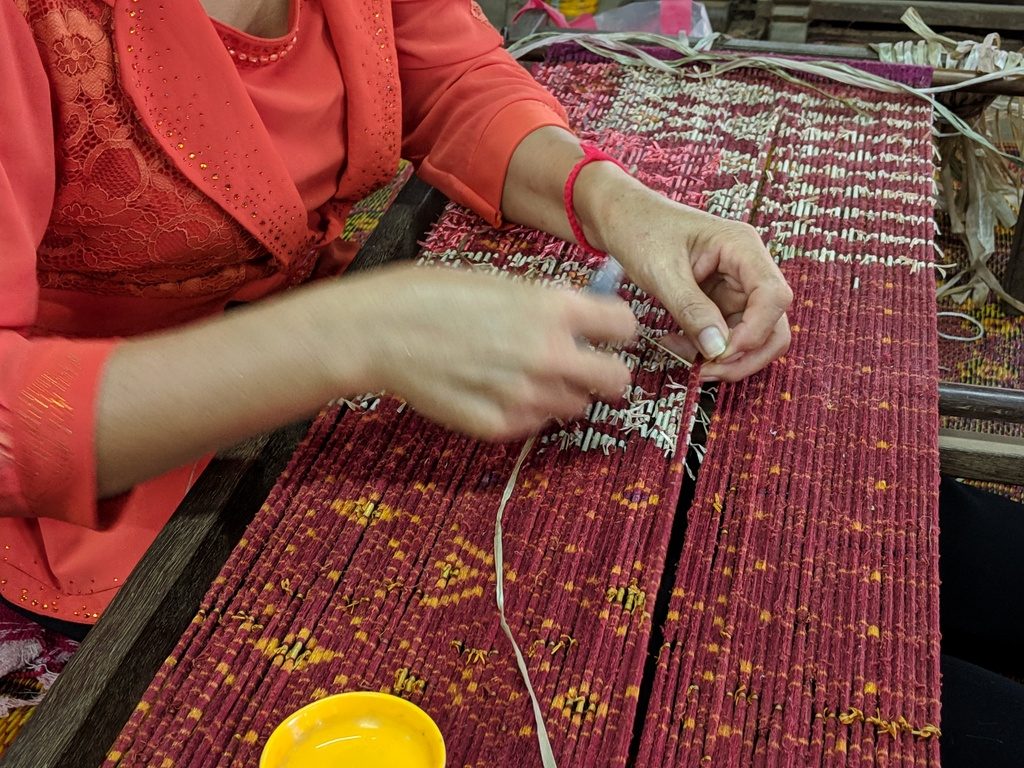 A close-up shot of a woman in a Cambodian village weaving a vibrant red and yellow mat on a traditional loom, with her hands actively working the threads