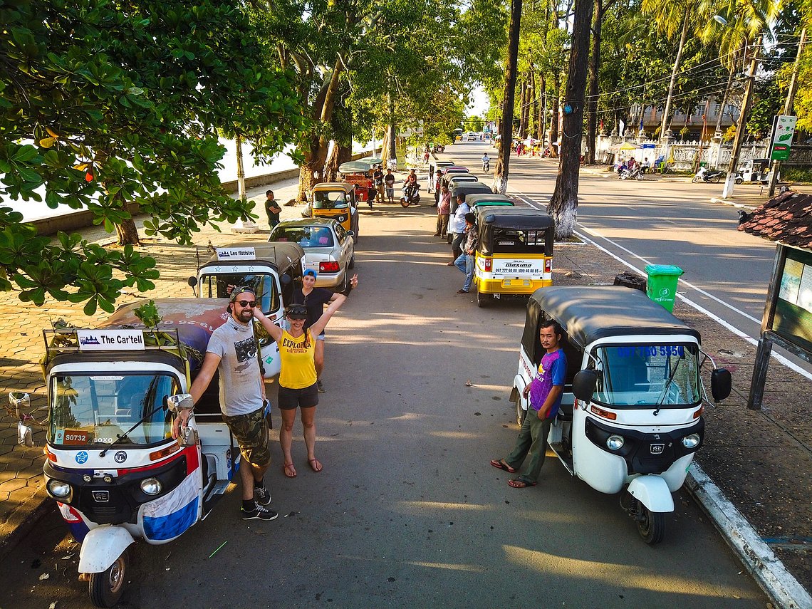 A row of tuk-tuks are parked along a tree-lined street in a Cambodian village