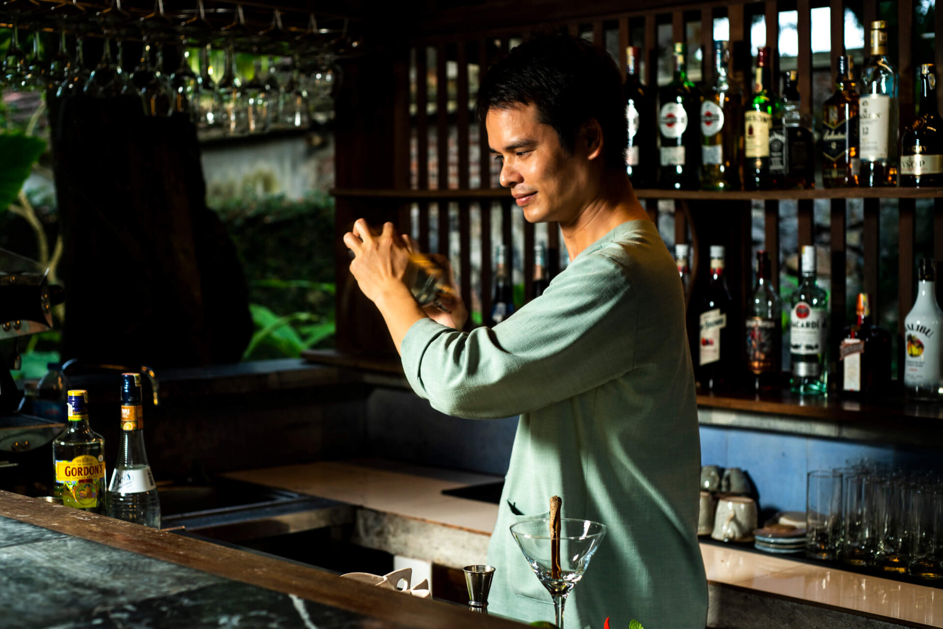 Bartender preparing cocktails at Longani Bar in Tam Coc Garden Resort with bottles, glasses, and warm atmosphere.