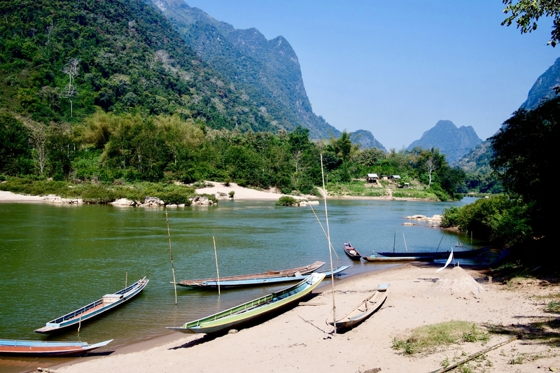 Traditional long-tail boats are pulled up onto the sandy bank of the Nam Ou River in Muang Ngoi, Northern Laos
