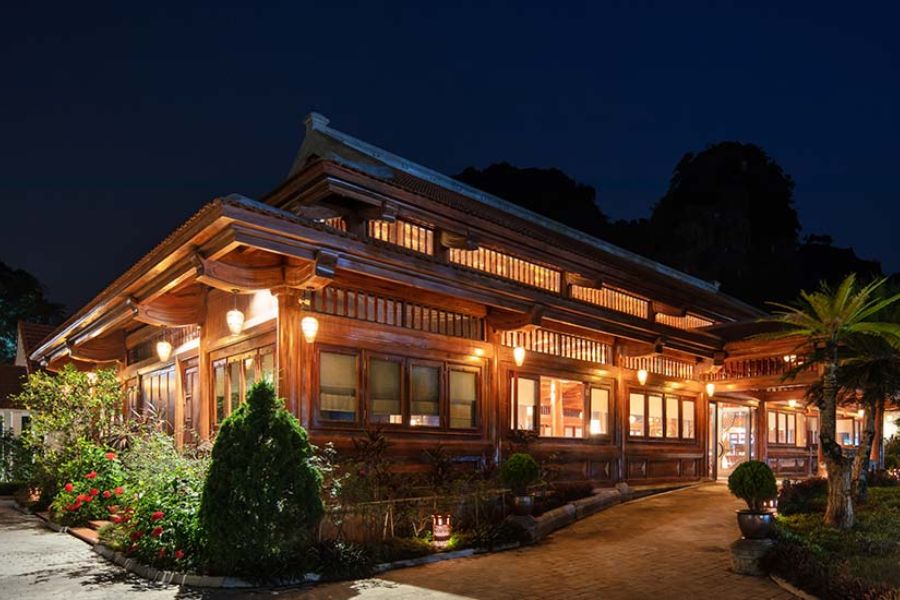 Nighttime exterior view of tam coc la montagne resort & spa restaurant building glowing with lights, surrounded by tropical gardens and mountains.