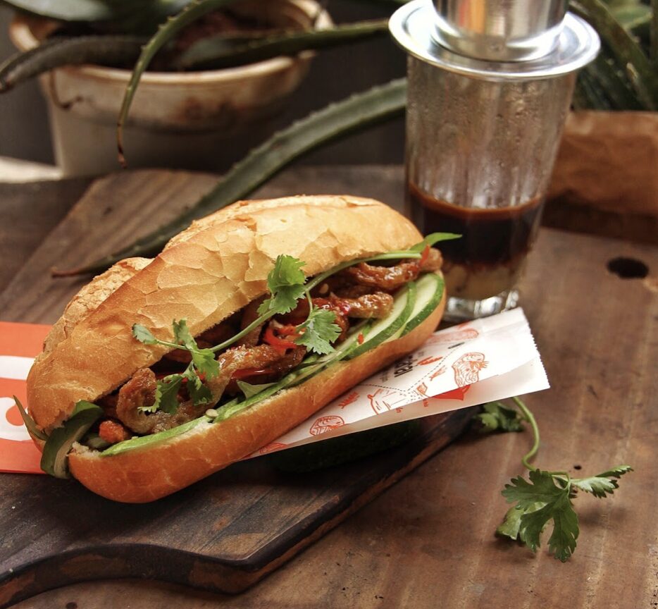 A crispy banh mi sandwich with grilled pork and herbs, a classic Vietnam street food served beside a traditional iced coffee.