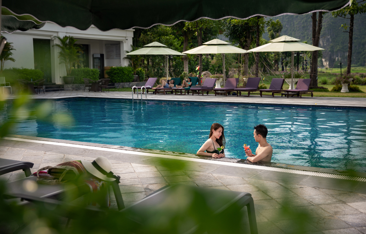Outdoor swimming pool at Ninh Binh Hidden Charm Hotel & Resort with guests relaxing under umbrellas and scenic mountain views.