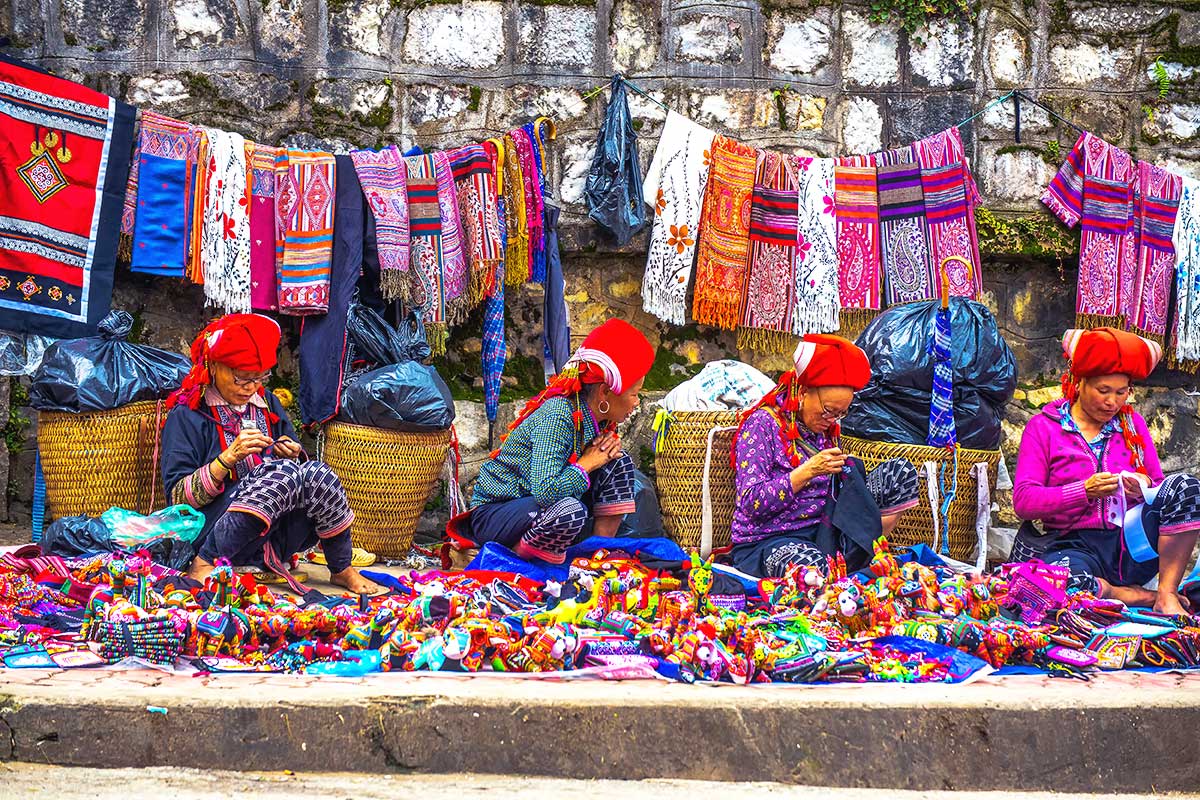 Four Red Dao women in traditional clothing and red head coverings sitting and hand-sewing crafts, a typical sight on a Sapa market tour