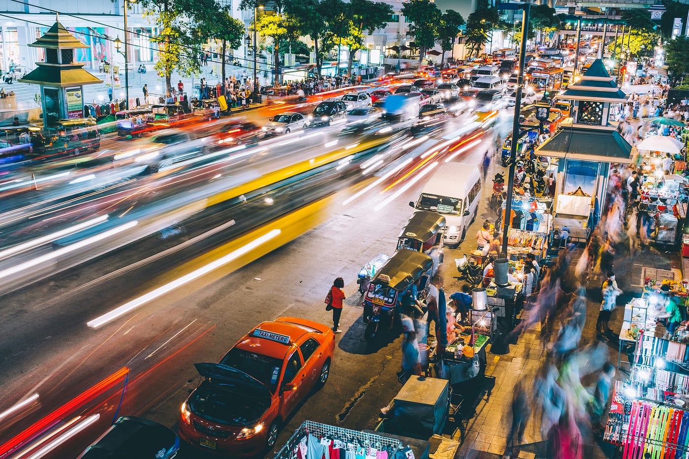 Vibrant long-exposure photo of a bustling street and night market in Bangkok, with streaks of light from fast-moving traffic