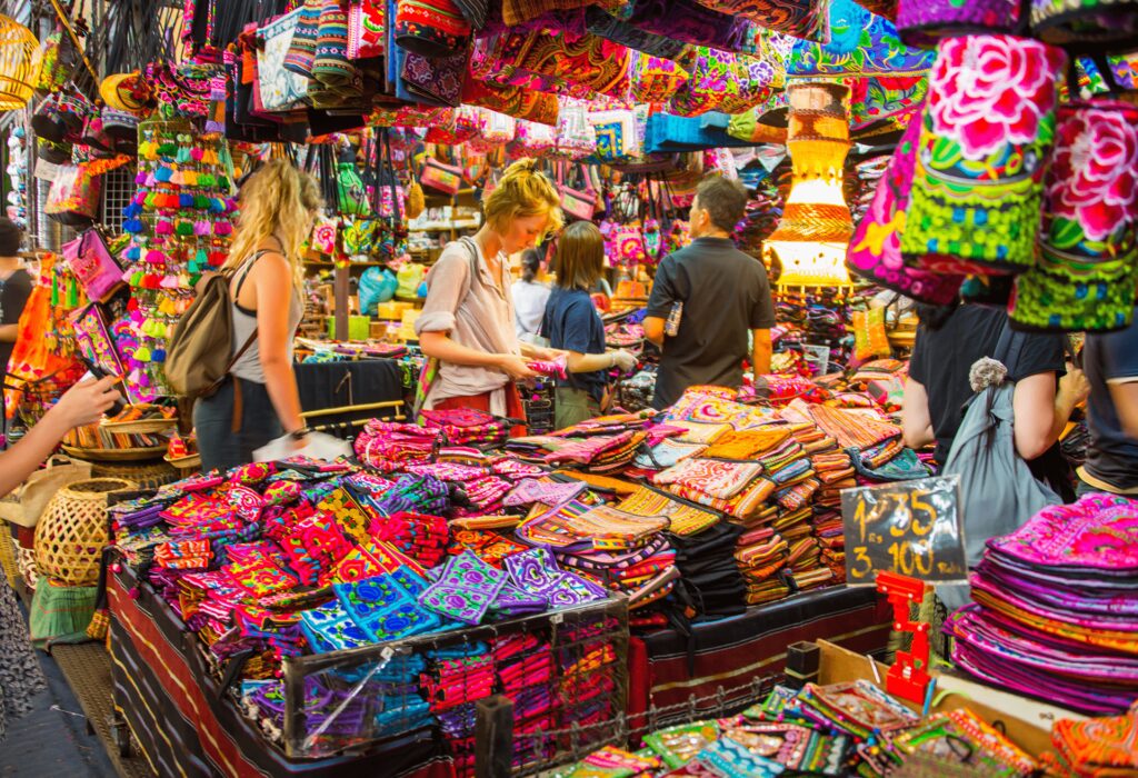 Tourists browsing brightly colored, patterned textiles, bags, and souvenirs stacked high at a crowded Bangkok night market stall