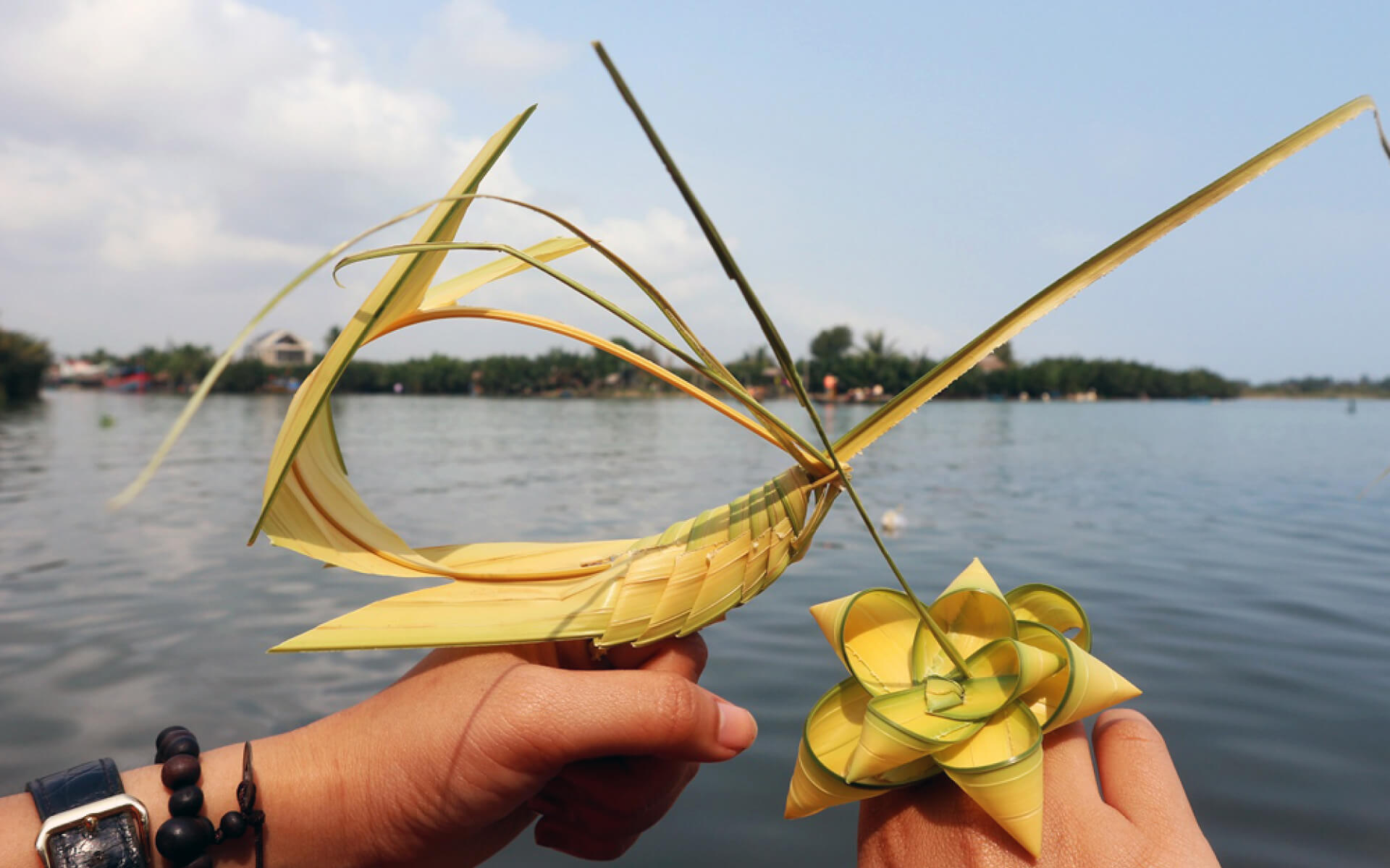 Handmade coconut-leaf souvenirs held by visitors beside peaceful riverside scenery at Bay Mau Coconut Forest.