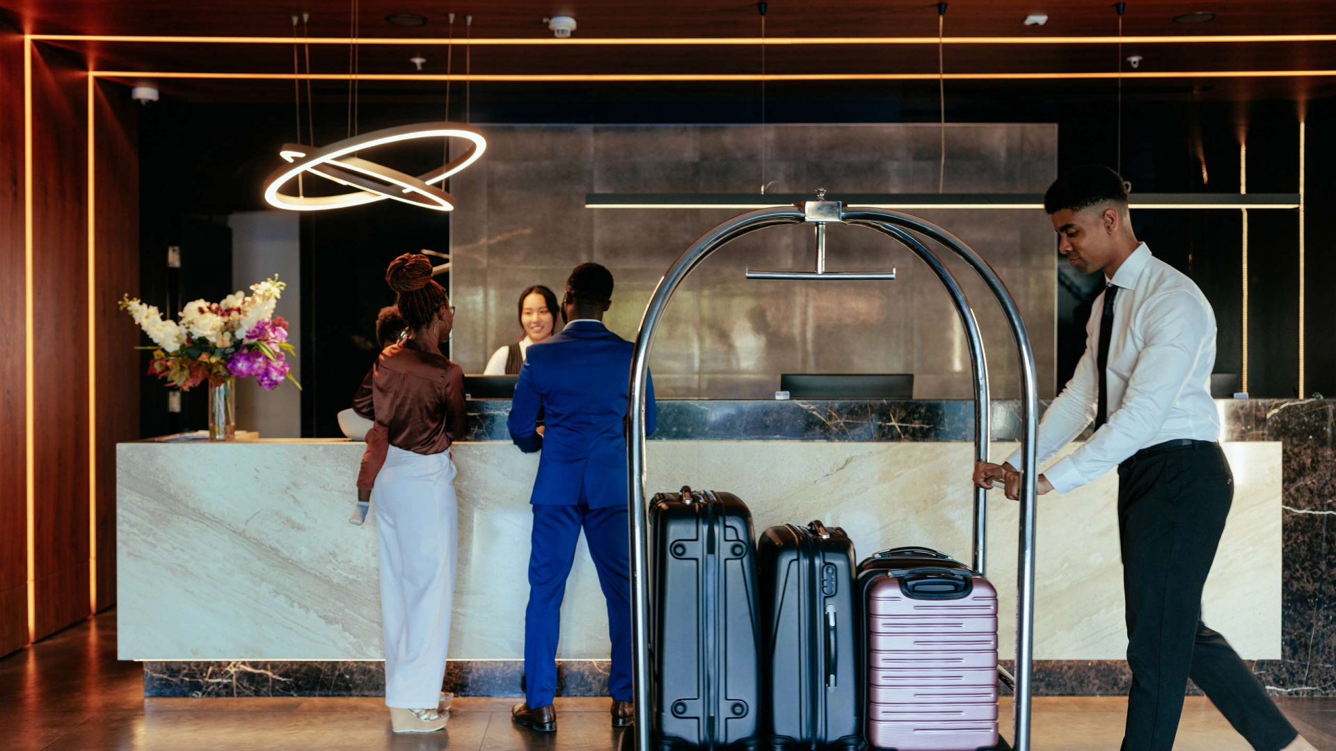 Hotel Accommodations: Guests checking in at a modern hotel reception desk while a bellhop moves luggage, illustrating luxury lodging services.