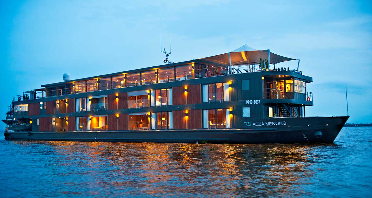 Luxury Mekong River Cruises: The Aqua Mekong cruise ship with multiple decks and illuminated cabins sailing on the river at dusk.