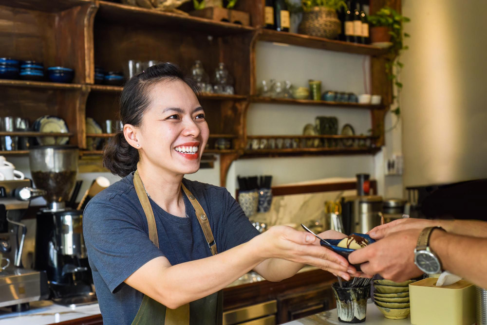 Restaurants and Cafés: Smiling female server handing a dessert to a customer across the counter, illustrating friendly service in a local café.