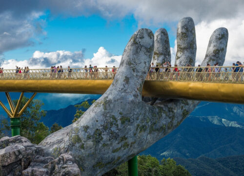 Tourists walking along Golden Bridge Hoi An supported by giant stone hands against dramatic mountain scenery.