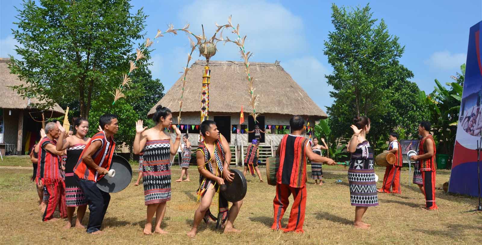 A group of performers from Vietnamese ethnic groups dancing in traditional attire in front of a thatched house, a cultural destination for hanoi day tours
