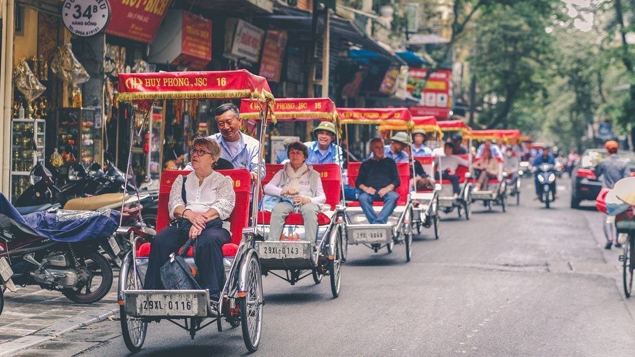 Tourists riding in a line of red-canopied cyclos down a busy street in Hanoi's Old Quarter, cyclo tour hanoi
