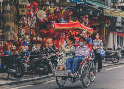 A cyclo carrying a family, passing a vibrant street stall selling balloons and decorations in Hanoi, one-day cyclo tour hanoi sightseeing