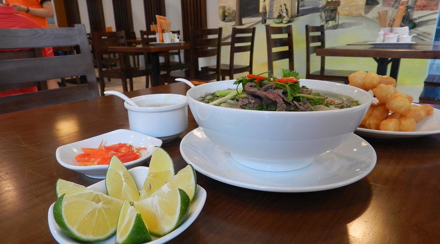 A close-up of a large white bowl of Pho with limes, chili, and fried dough sticks, representing a culinary stop on hanoi day tours