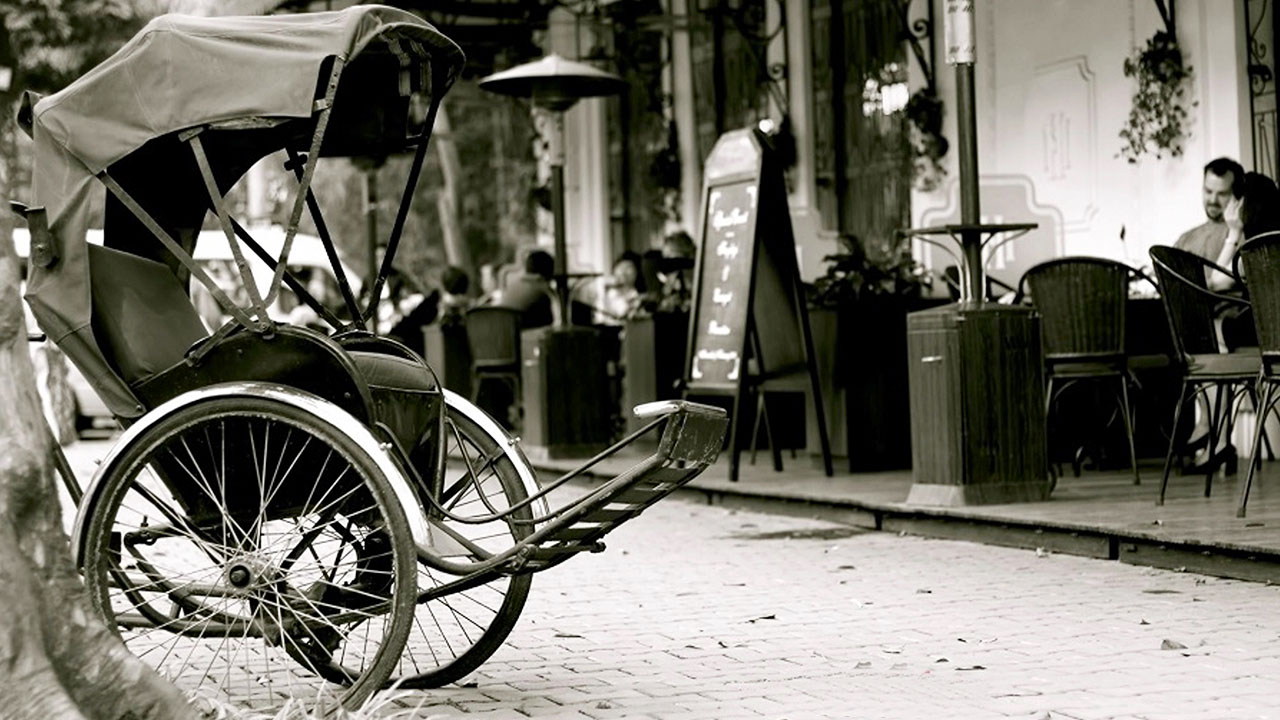A vintage cyclo parked on a cobblestone street near a cafe, symbolizing the historical and cultural significance of the cyclo tour hanoi