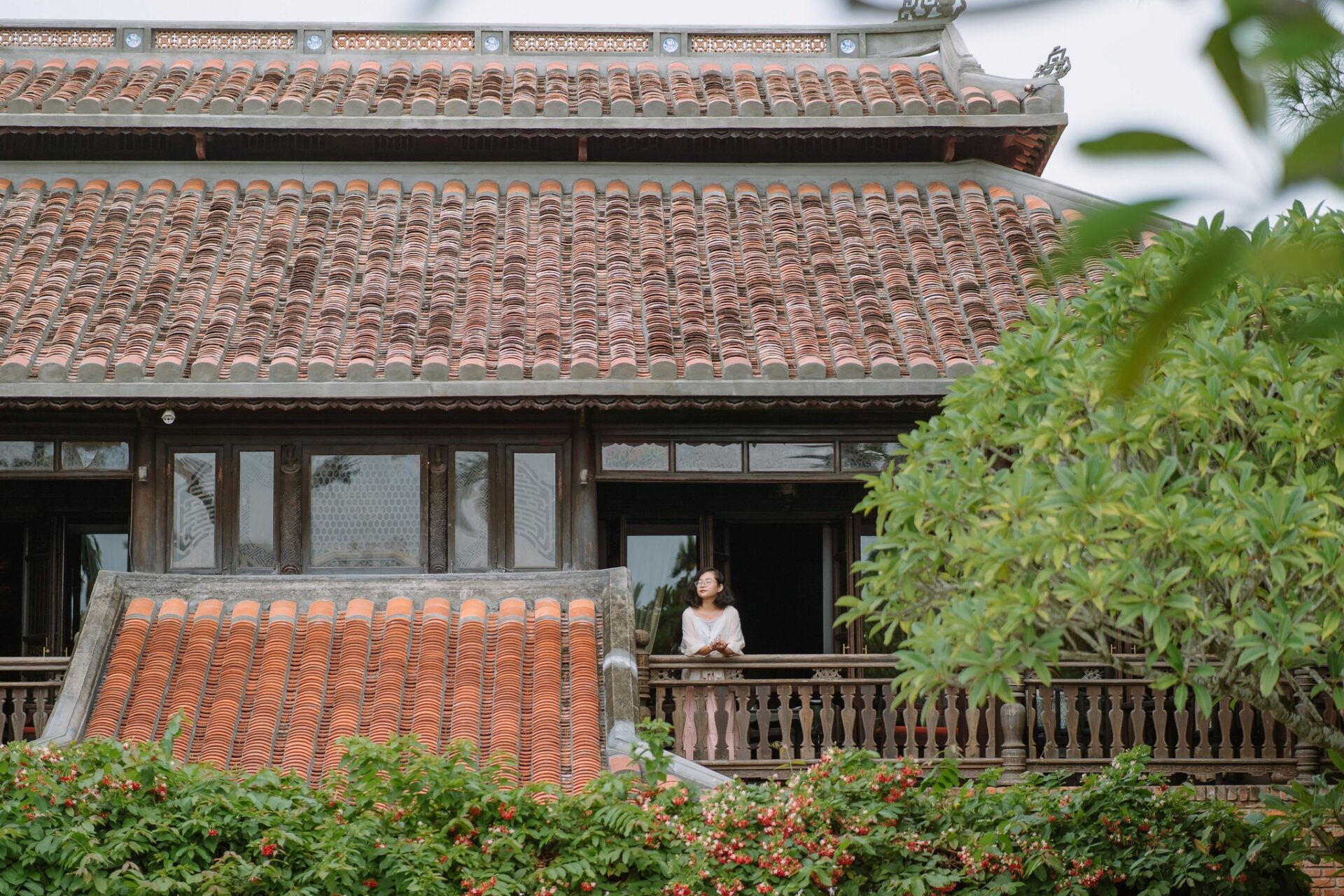Traditional tiled-roof architecture at Ancient Hue Garden Houses with lush greenery and a guest enjoying the tranquil view from the balcony.