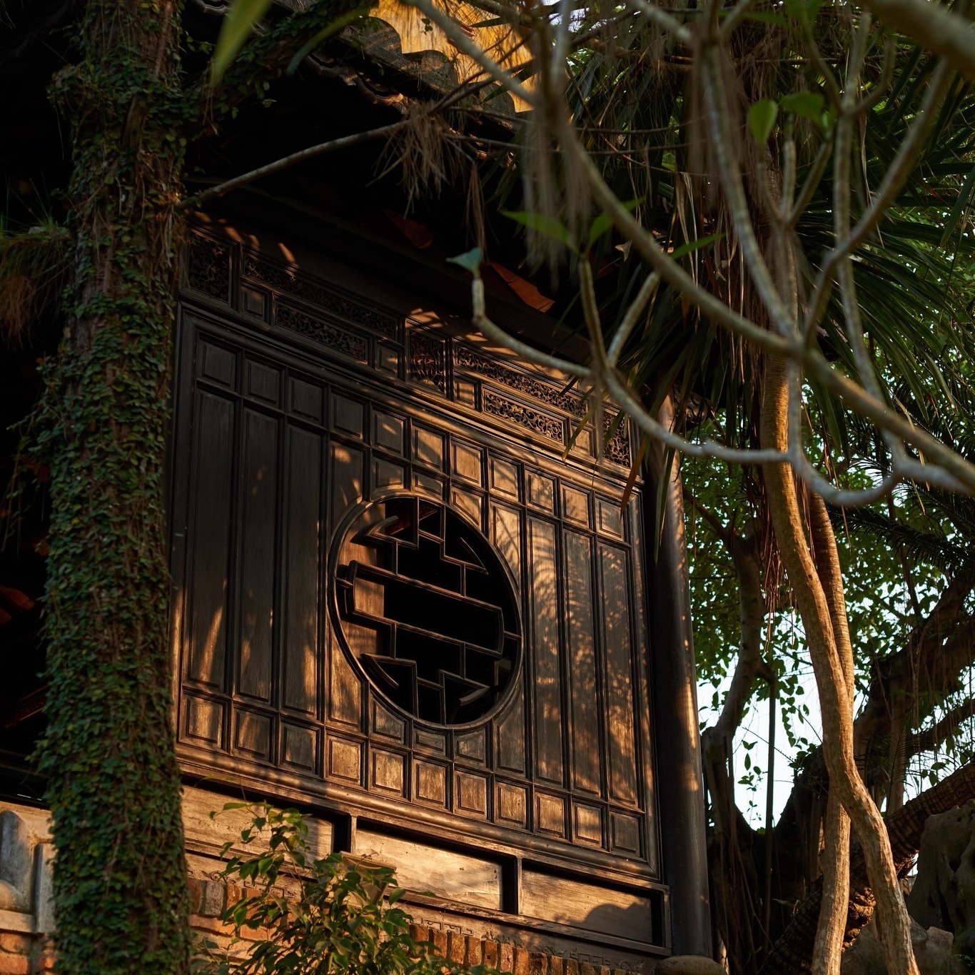 Traditional wooden architecture at Ancient Hue Garden Houses with circular window, intricate carvings, and warm sunlight filtering through lush tropical trees.