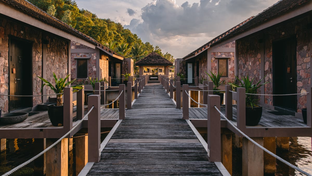 Wooden walkway connecting over-water villas at Vedana Lagoon Resort Vietnam during sunset, featuring rustic stone architecture.