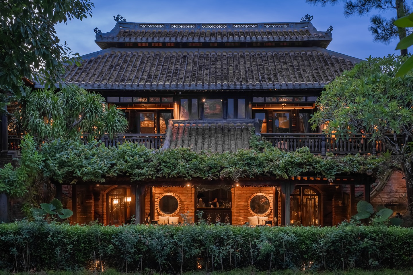 Traditional Vietnamese architecture at Ancient Hue Garden Houses, featuring tiled roofs, wooden pillars, and lush gardens illuminated at dusk.