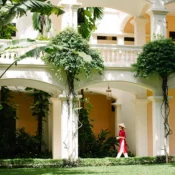 A Vietnamese woman in ao dai walks beneath colonial arches surrounded by tropical gardens at Anantara Hoi An Resort.