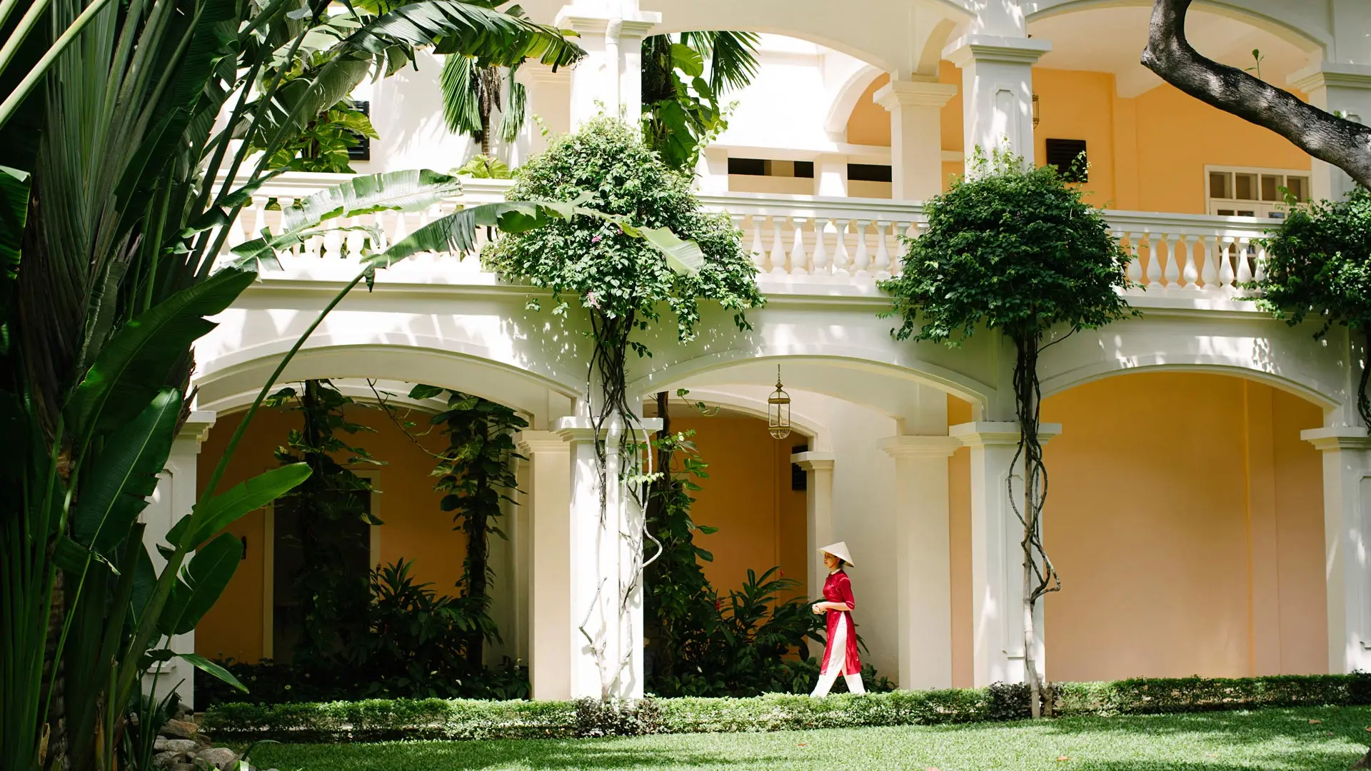 A Vietnamese woman in ao dai walks beneath colonial arches surrounded by tropical gardens at Anantara Hoi An Resort.