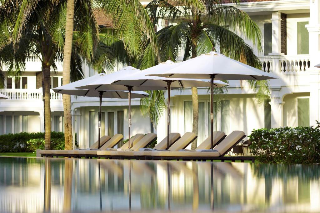 Sun loungers and white umbrellas reflecting in the water beside palm trees at Boutique Hoi An Resort’s serene pool area.