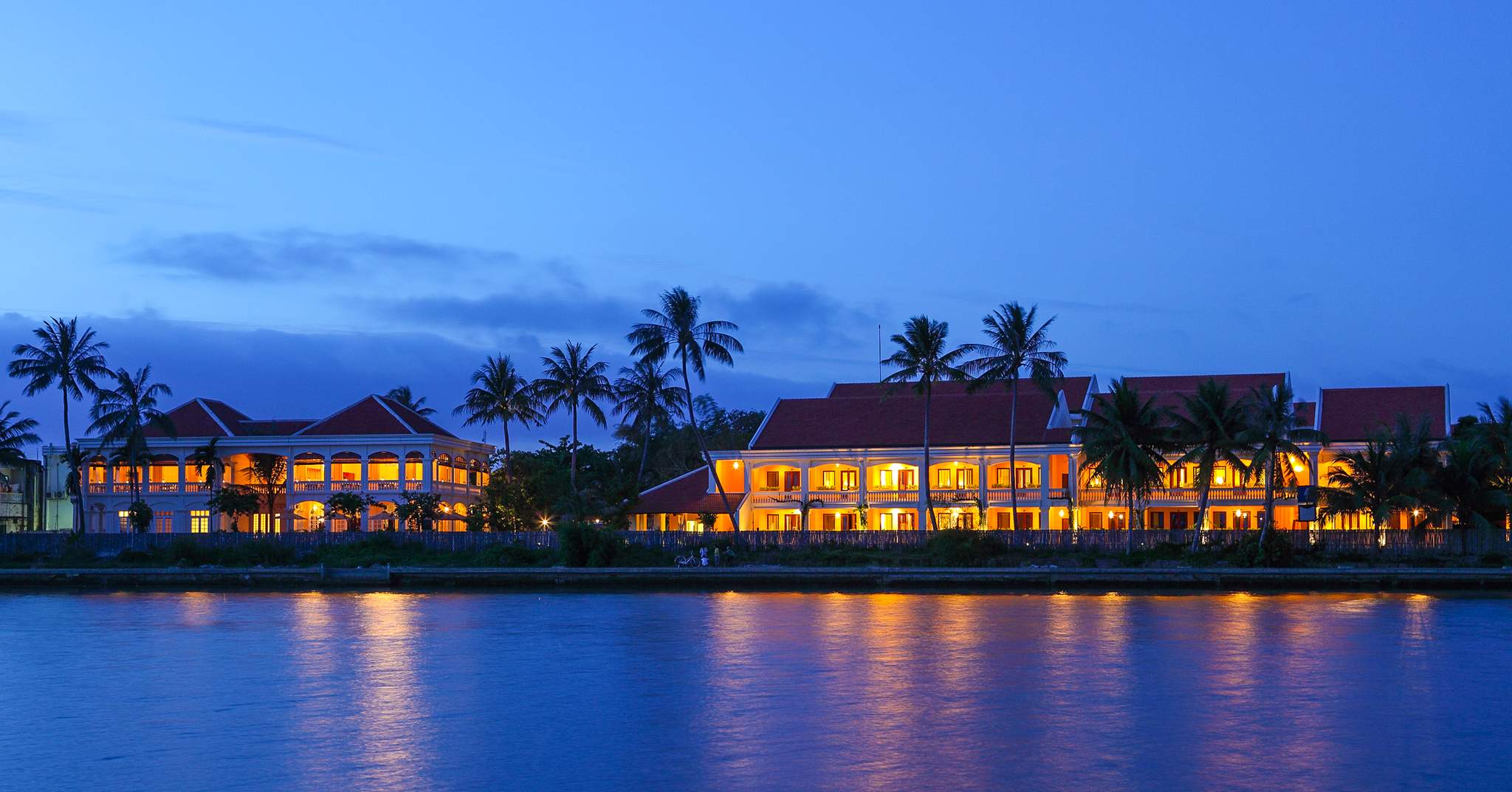 Twilight view of Anantara Hoi An Resort glowing beside the Thu Bon River with palm trees reflecting lights.