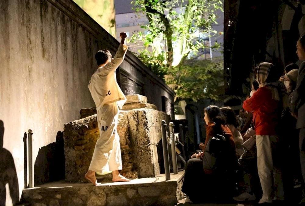 Night Tour: Exploring the 'Hell' of Hanoi Hilton Prison: Actor in a white prison uniform (M.C.) reenacting a scene with a raised fist for tourists during the popular night tour.
