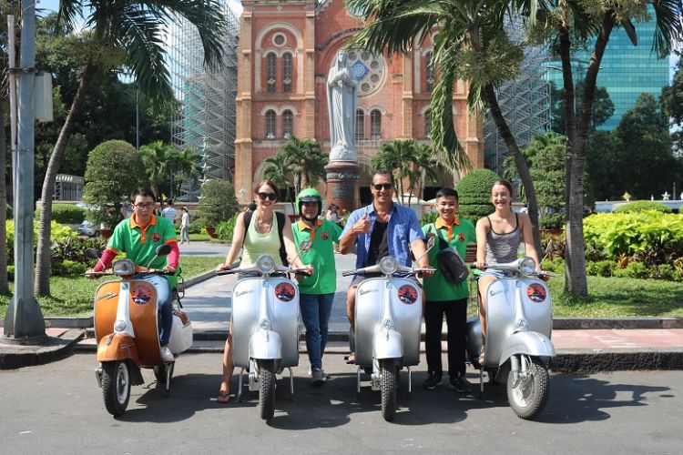 A group of smiling travelers wearing helmets and sitting on red scooters, ready to begin their motorbike adventure as part of a Ho Chi Minh City tour